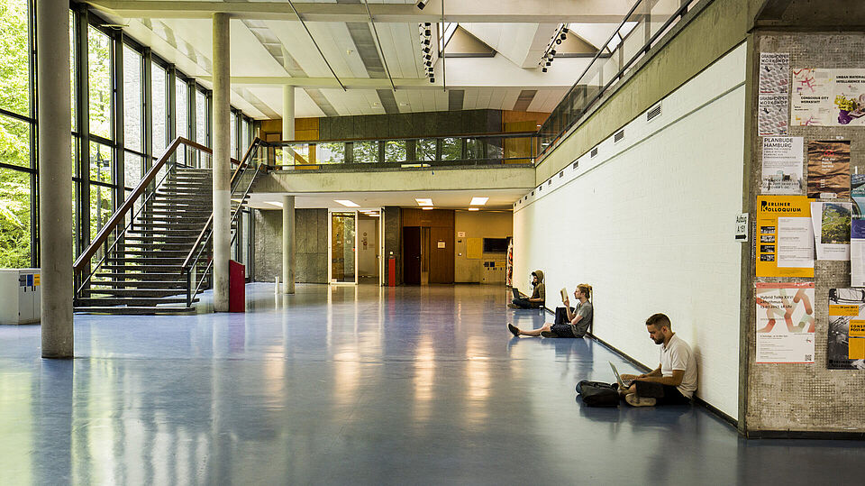 Erdgeschoss im Inneren des Foyers im Architekturgebäude, Studierende sitzen gegenüber der Glasfassade an der Wand gelehnt und studieren 