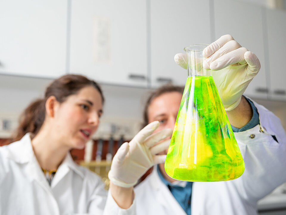 Zwei wissenschaftliche Mitarbeiter betrachten einen Laborflasche mit Fluorescin, das in wässriger Lösung unter ultraviolettem Licht grün leuchtet