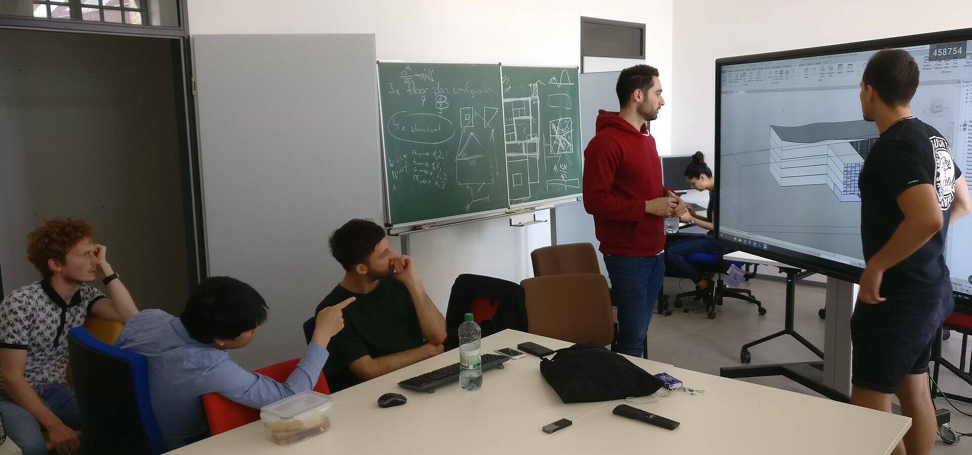 Sechs Studierende in einem Seminarraum stehend und sitzend im Gespräch über einen Gebäudeplan, der auf einem großen Smartboard angezeigt wird