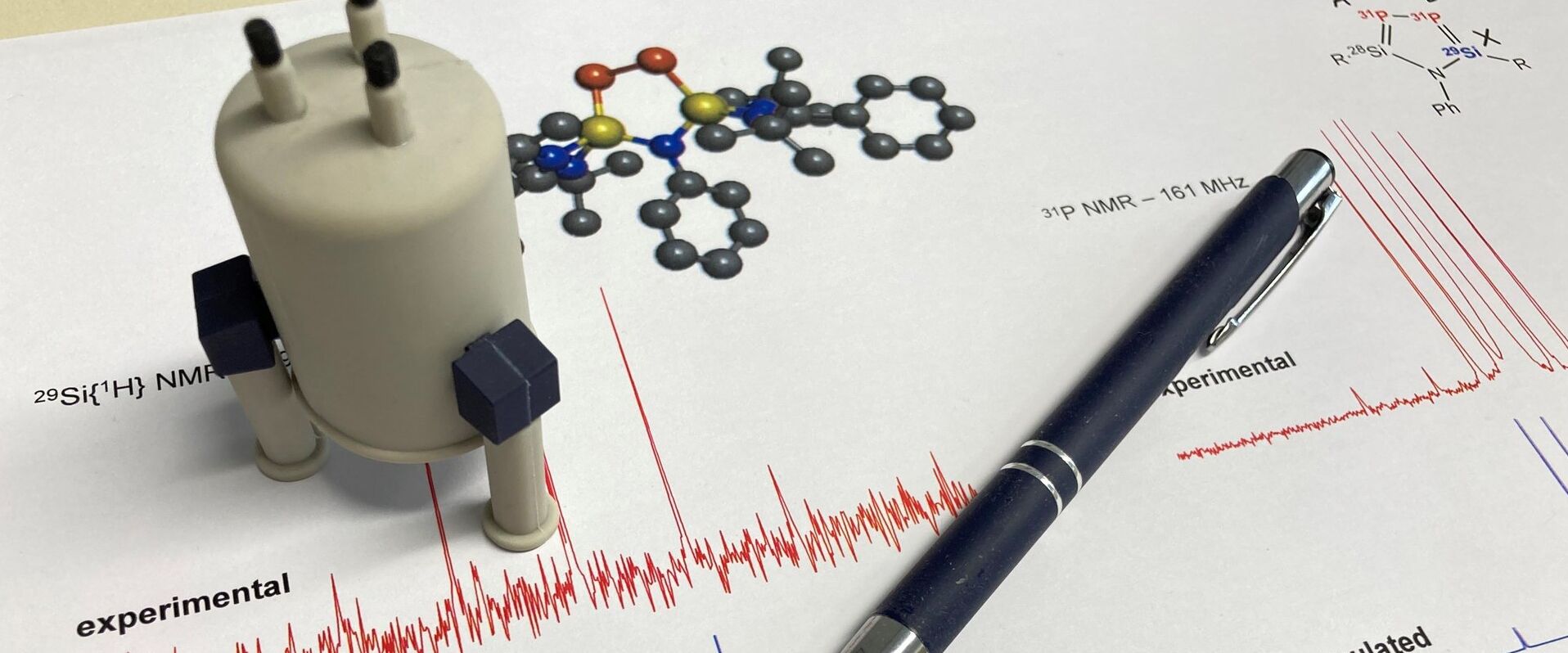 Ein ausgedrucktes NMR-Spektrum mit Molekülstruktur, darauf ein Stift und Miniatur NMR-Gerät