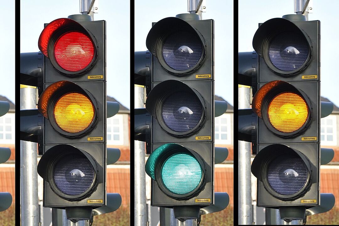 Vier Verkehrsampeln dicht nebeneinander in der Nahaufnahme. Sie zeigen unterschiedliche Lichtsignale an. Von links nach rechts ist die erste Ampel rot, die Zweite rot und gelb, die Dritte grün und die Vierte gelb.