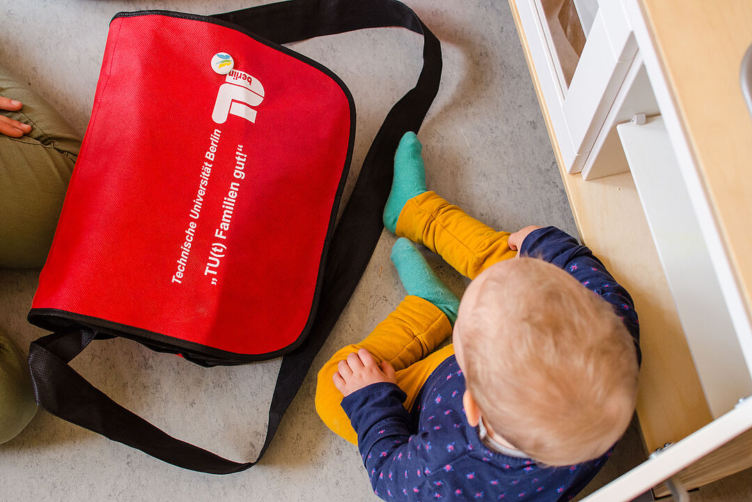 view of a toddler from above, sitting next to a bag, the TU logo and the inscription "Tu(t) Familien gut" (good for families) can be seen on the bag