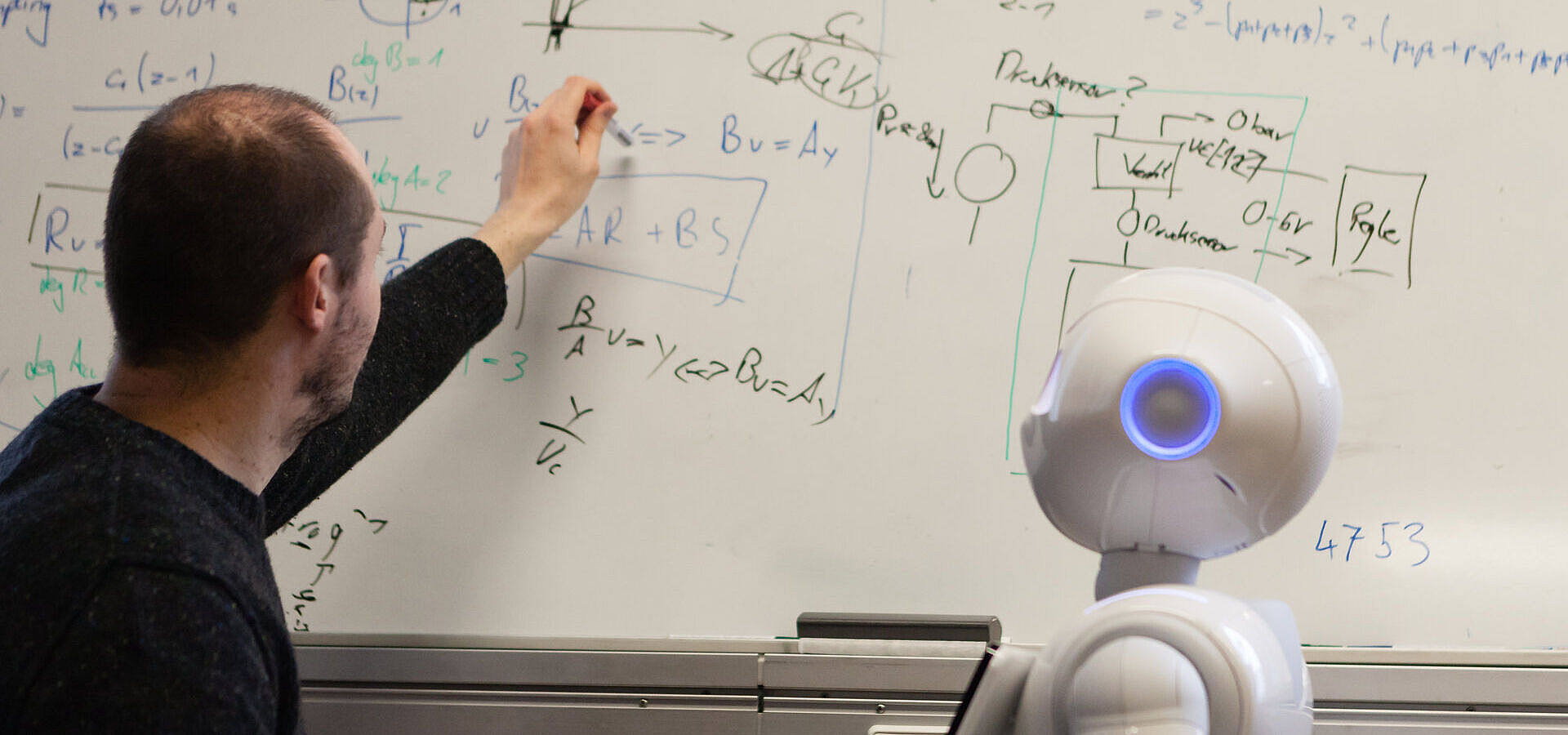 Wissenschaftler der Technischen Universität Berlin bei der Interaktion mit dem humanoiden Roboter Pepper, vor einer Tafel stehend etwas erläuternd 