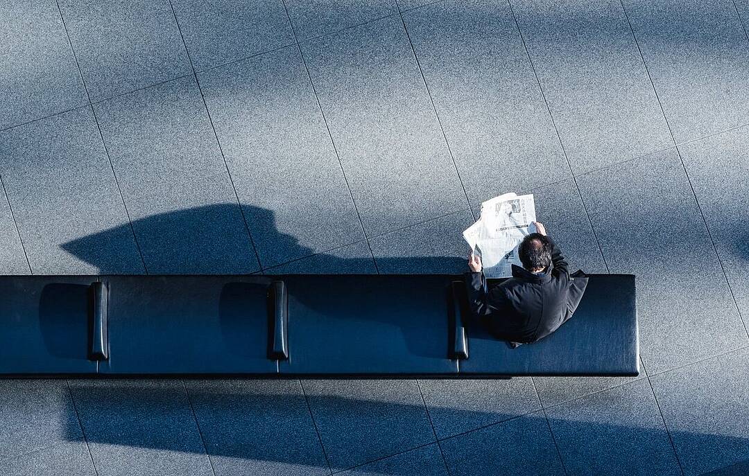 Vogelperspektive: Person sitzt auf einer schwarzen Bank und liest Zeitung