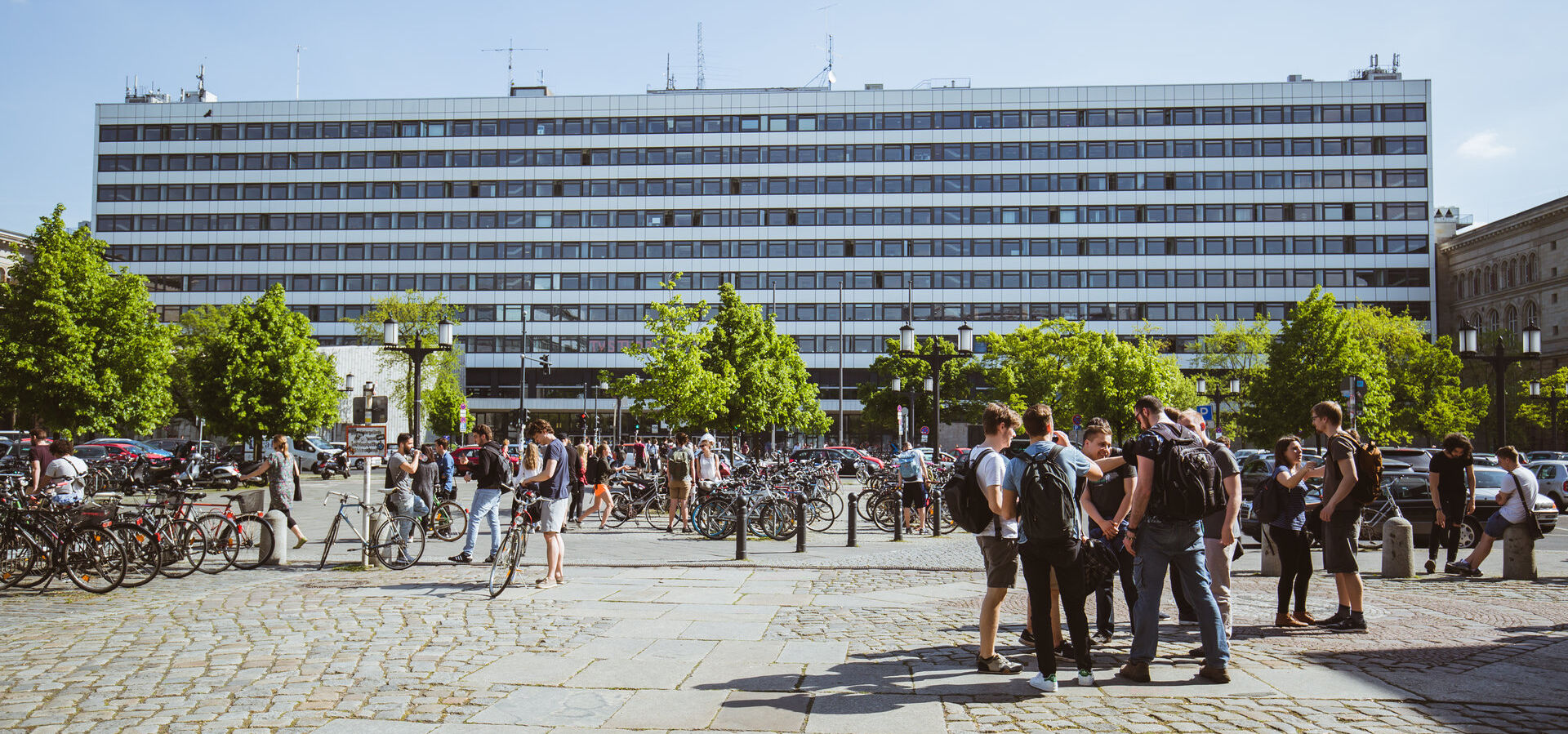 Blick auf das Hauptgebäude über die Straße des 17. Juni bei Sonnenschein, Studierende stehen in Gruppen