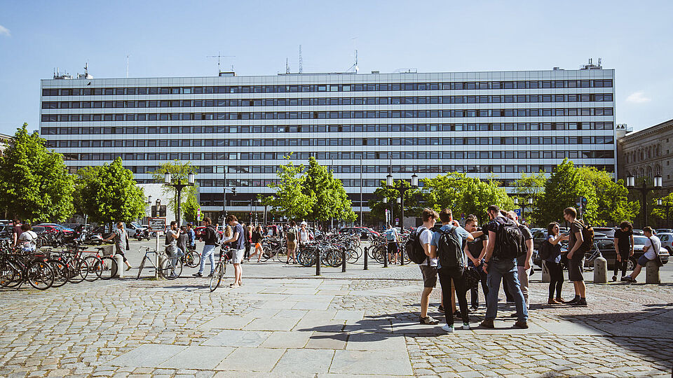Blick auf das Hauptgebäude über die Straße des 17. Juni bei Sonnenschein, Studierende stehen in Gruppen