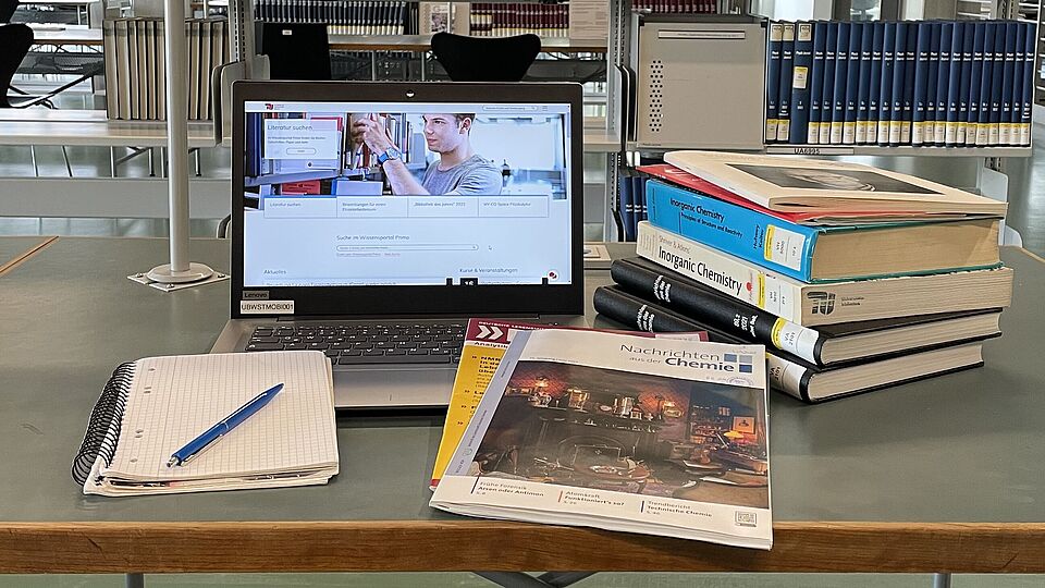 Ein Arbeitsplatz in der Bibliothek mit Laptop und einem Stapel aus Büchern und Zeitschriften