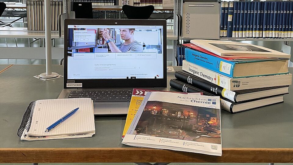 Ein Arbeitsplatz in der Bibliothek mit Laptop und einem Stapel aus Büchern und Zeitschriften