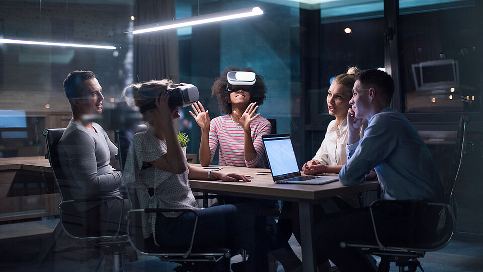 Multiethnisches Business-Team mit Virtual-Reality-Headset in Nachtbüro Treffen Entwickler mit Virtual-Reality-Simulator rund um Tisch in Kreativbüro.