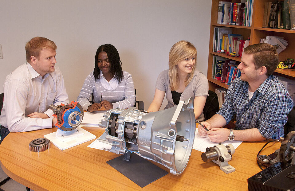 Vier Studierende in einem Gruppengespräch. Auf dem Tisch finden sich verschiedene Objekte aus dem Bereich Maschinenbau