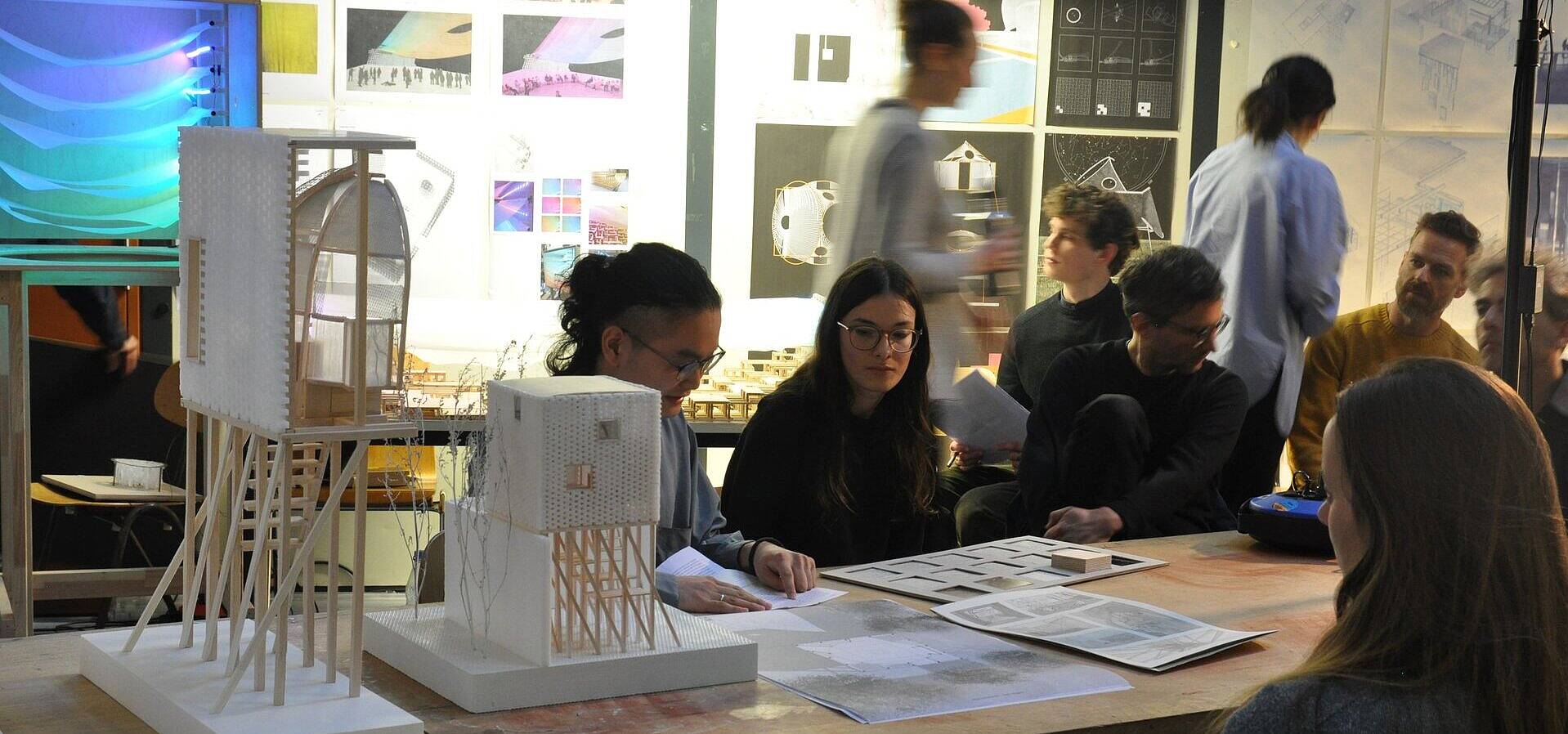 Neun junge Personen in einem Raum voller architektonischer Objekte und Installationen, 3 Personen besprechen einen Plan, die anderen bewegen sich im Raum oder sitzen