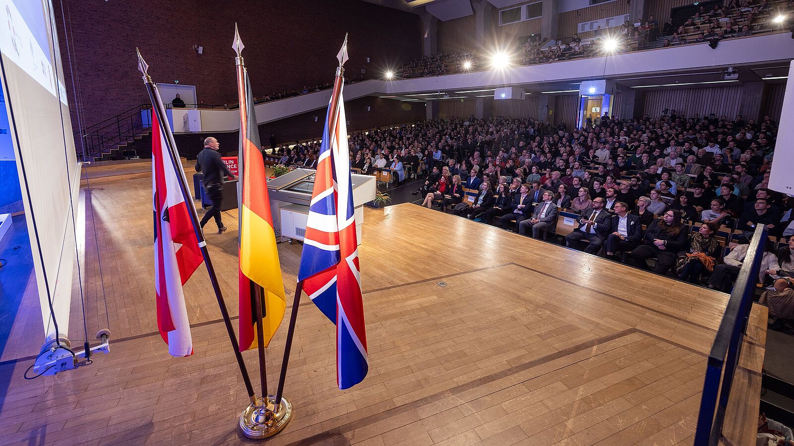 Blick von der Bühne ins Audimax. Im Vordergrund die Flaggen Großbritanniens, Deutschlands und Berlins.