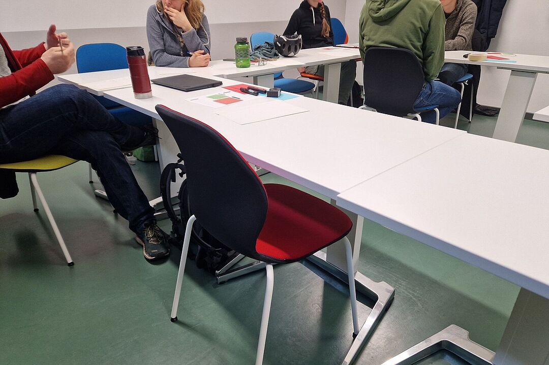 In einem kleinen Schulungsraum sind Tische und Stühle zu sehen. An einigen Plätzen sind Workshop Teilnehmer zu erkennen aber ohne Gesicht.