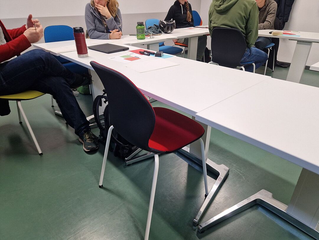 Tables and chairs can be seen in a small training room. In some places workshop participants can be seen but without their faces.