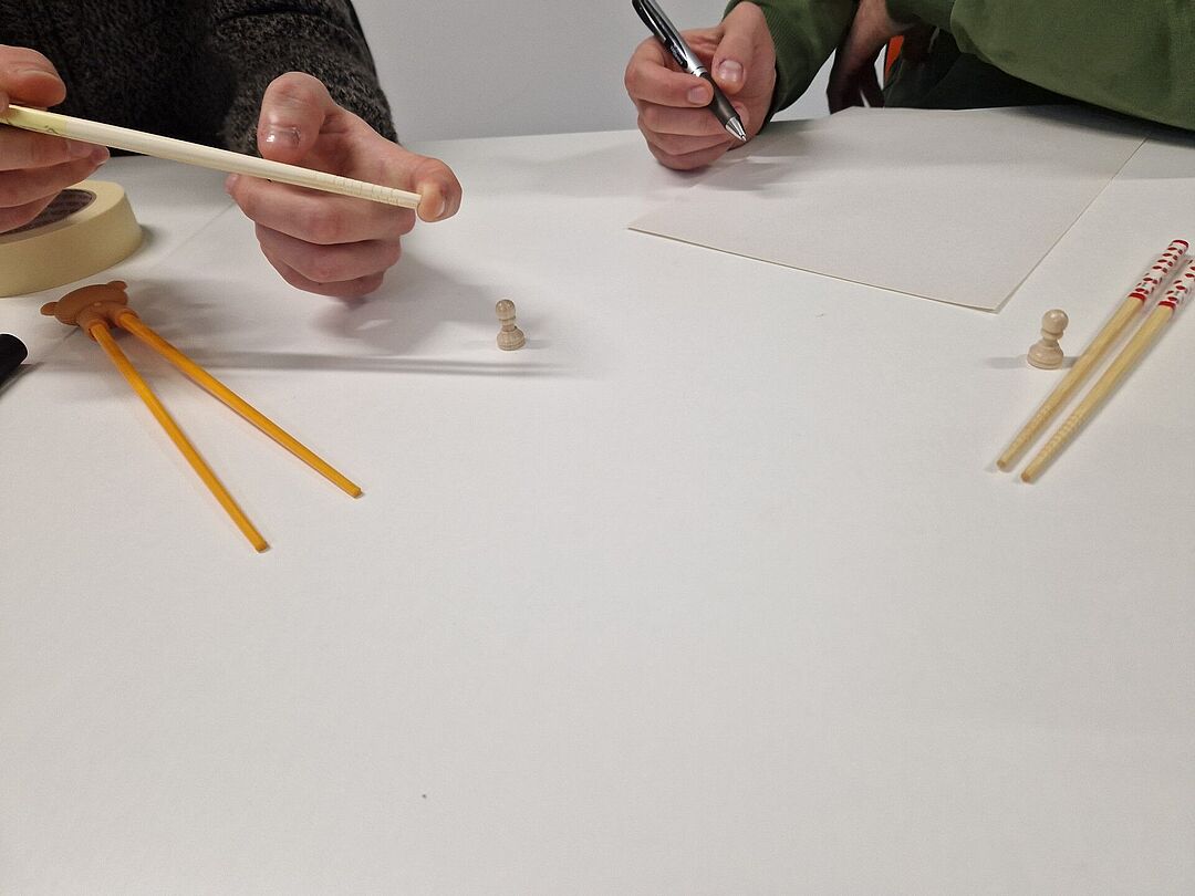 Only the hands of 2 people can be seen at a table. A person holds a pair of chopsticks in their hands. The other person has a blank sheet of paper in front of them and is holding a pen.
