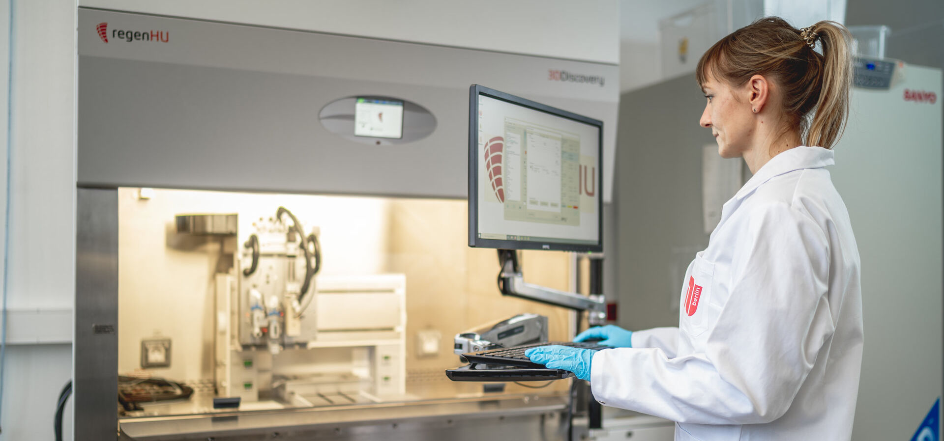 Eine junge Wissenschaftlerin steht an einem Monitor in einem Labor des Fachgebietes Angewandte Biochemie und schaut auf einen Bildschirm, dahinter Laborgeräte