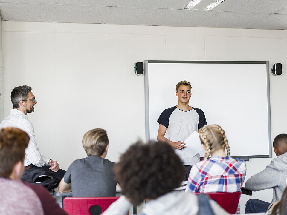 Klassenzimmersituation in der ein junger Mann vor einer Schulklasse steht und spricht, während ein älterer Lehrer den Unterricht beobachtet