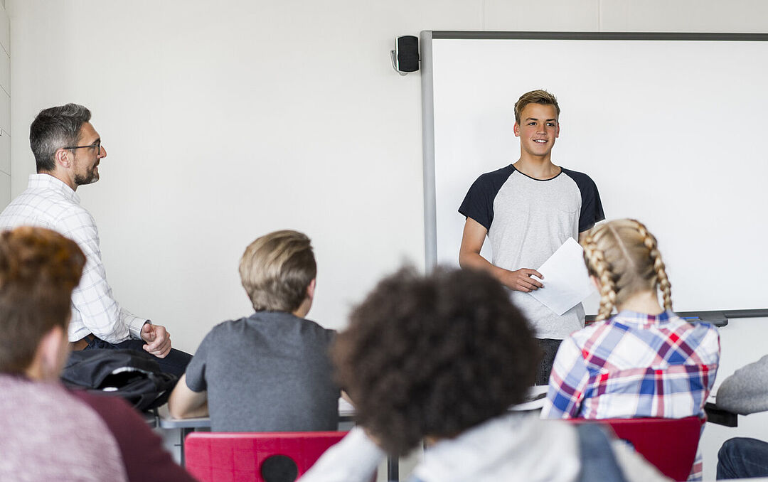 Klassenzimmersituation in der ein junger Mann vor einer Schulklasse steht und spricht, während ein älterer Lehrer den Unterricht beobachtet