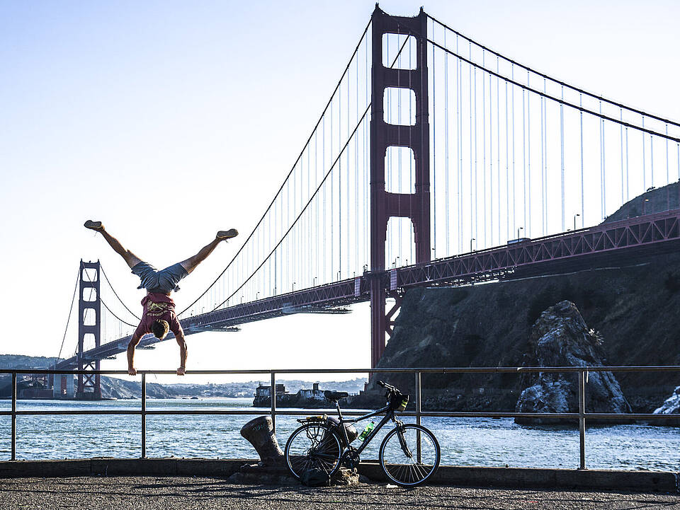 Yannic Scholtyssek macht einen Handstand am Ufer vor der Golden Gate Bridge in San Francisco