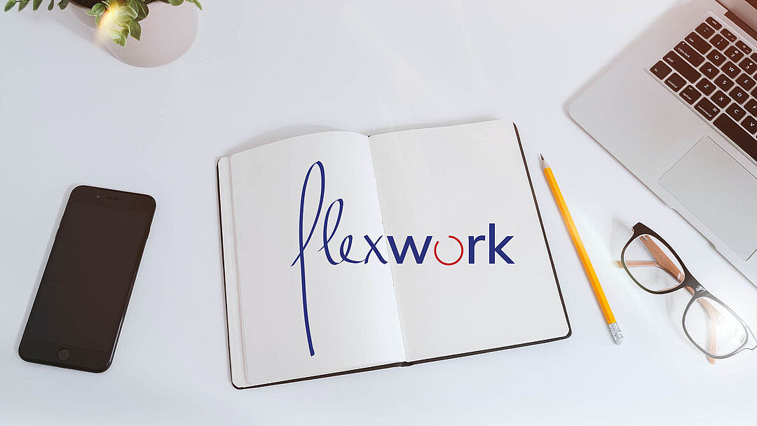 Ein aufgeschlagenes Notitzbuch mit Schriftzug "flexwork" über beide Seiten; daneben liegen ein Handy, ein Bleistift, eine Brille und ein Laptop (nur halb zu sehen)