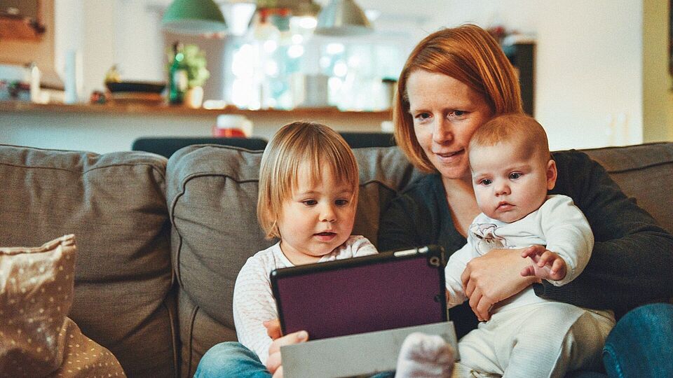 Eine Frau hat zwei Babys auf dem Schoß und sie schauen sich etwas auf dem Tablet an. 
