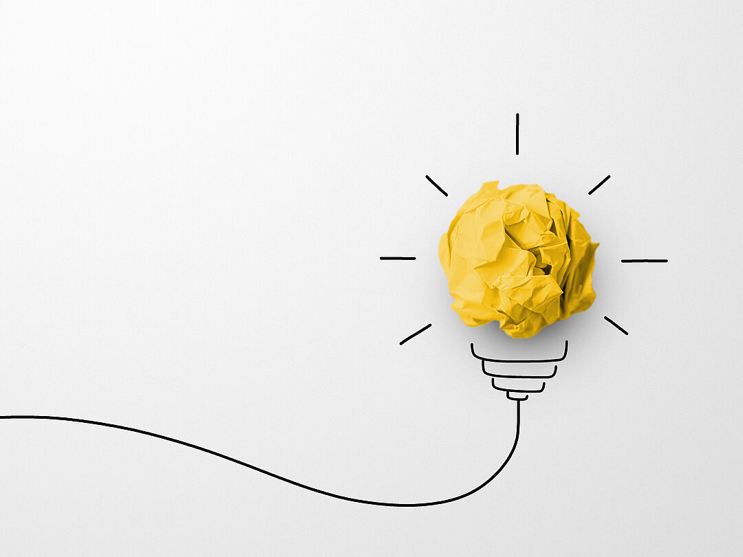 a crumpled piece of yellow paper on a light background, around the piece of paper a cable socket and rays have been added with a black pen, so that the impression of a light bulb is created