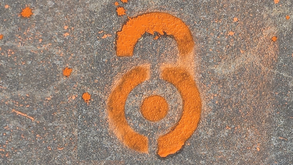 Das Foto zeigt das Bodengraffiti des OA-Logos mit orangefarbiger Sprühkreide auf einem dunkelgrauen Betonboden.