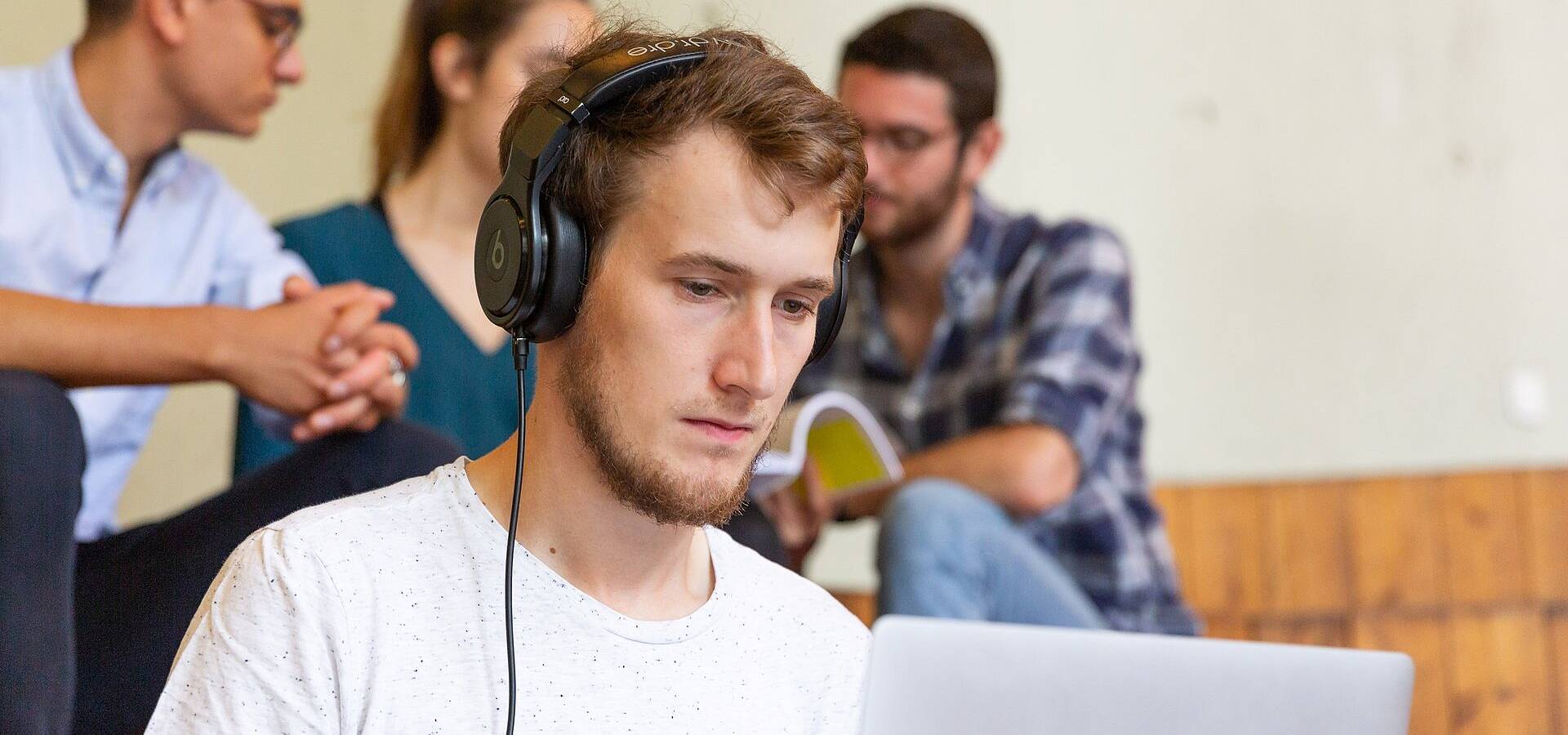 Studierende in einem Gebäudeteil der Technischen Universität Berlin sitzend und im Gespräch auf Stufen, im Vordergrund sitzt einer junger Studierender mit Kopfhörern, der konzentriert mit einem Laptop arbeitet