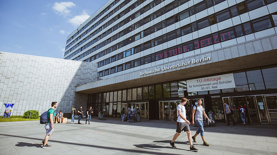 Haupteingang mit angeschnittenem Audimax und Hauptgebäude, Studierende betreten und verlassen das Gebäude
