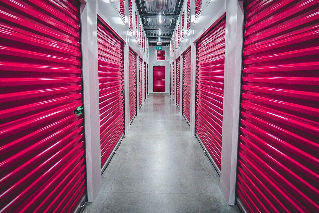 Ein Gang, wo links und rechts geschlossene pinkfarbenen Rolltore zu Lagerräumen zu sehen sind.