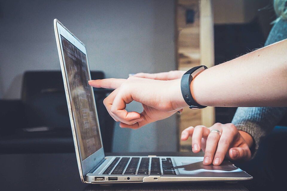 Ein Laptop steht aufgeklappt auf einem Tisch. Eine Hand zeigt auf den Bildschirm und eine andere Hand benutzt das Mousepad. Beide Hände sind von unterschiedlichen Personen.