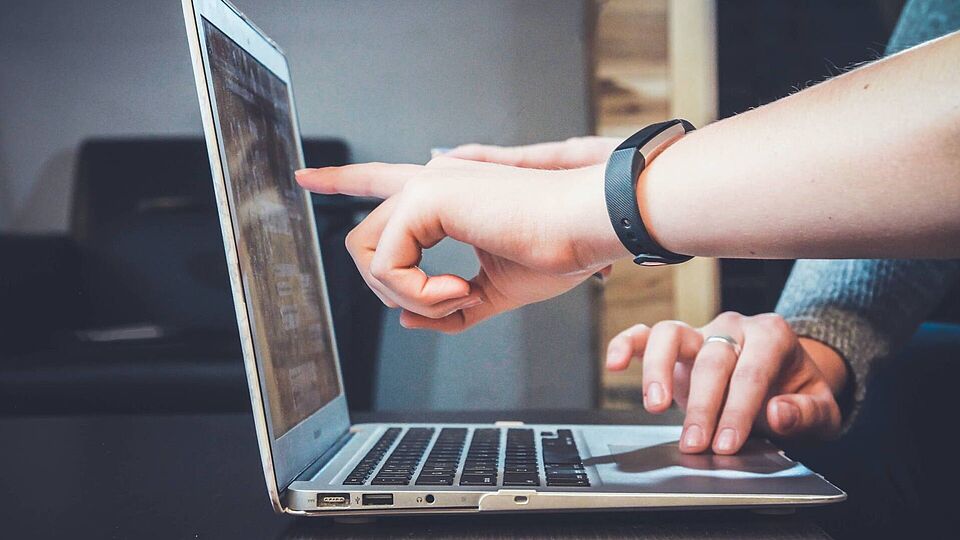 A laptop is open on a table. One hand is pointing at the screen and another hand is using the mousepad. Both hands are from different people.